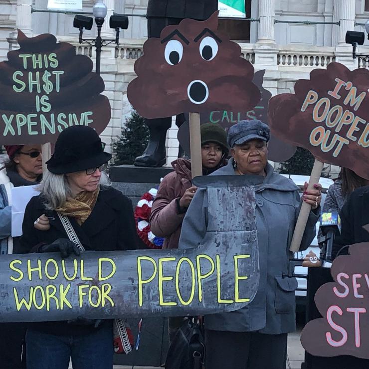 A crowd of people rallying outside of Baltimore City Hall, with signs like "Pipes should work for people" and "sewage stinks"